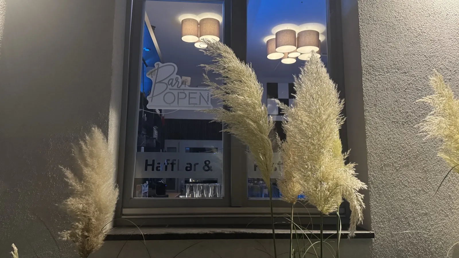 Beleuchtetes Fenster der Haffbar im Alten Bahnhof mit Schild „Bar open“ und Pampasgras davor